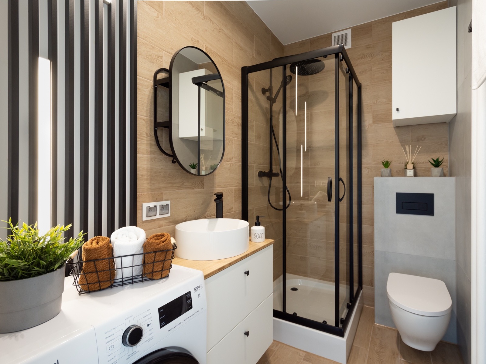 Nowoczesna łazienka z kabiną prysznicową w czarnej ramie, umywalką nablatową na białej szafce z drewnianym blatem i pralką pod blatem, uzupełniona o owalne lustro i drewnopodobne płytki.