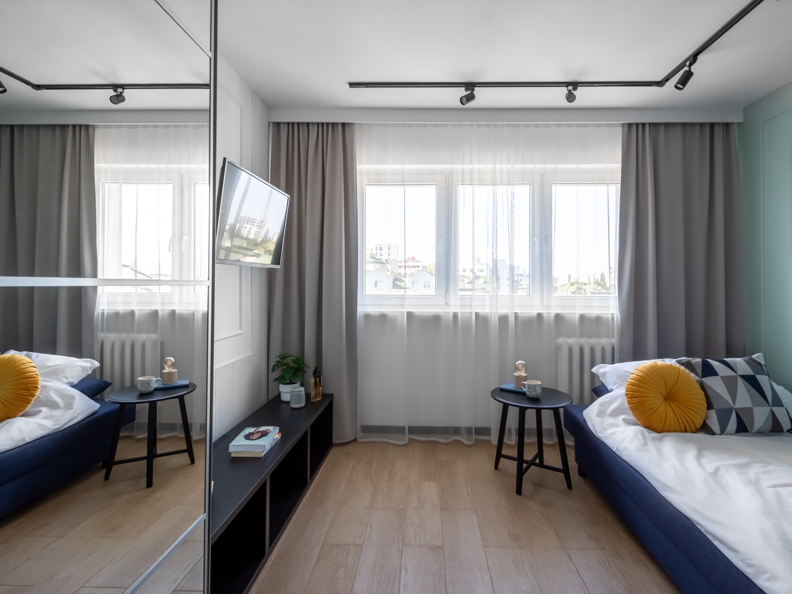 Nowoczesna sypialnia z łóżkiem w niebieskiej ramie, drewnianą podłogą, czarnymi stolikami i półką, oświetleniem szynowym i dużym lustrem w szafie, odbijającym część pokoju.