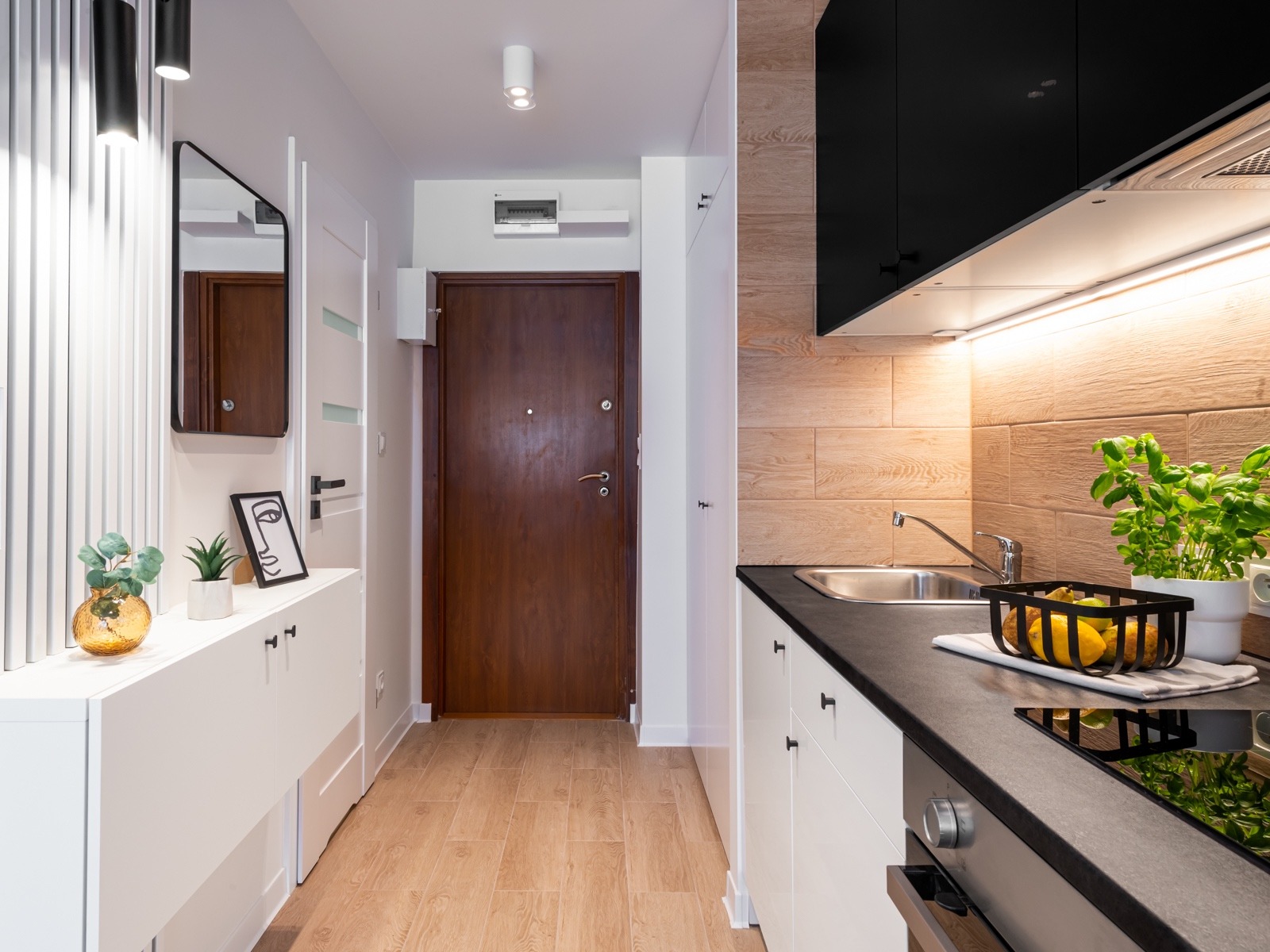 Nowoczesne wykończenie wnętrza mieszkania: drewnopodobne płytki na podłodze i ścianie, białe szafki z czarnymi uchwytami, czarne szafki wiszące w kuchni, lustro w przedpokoju.