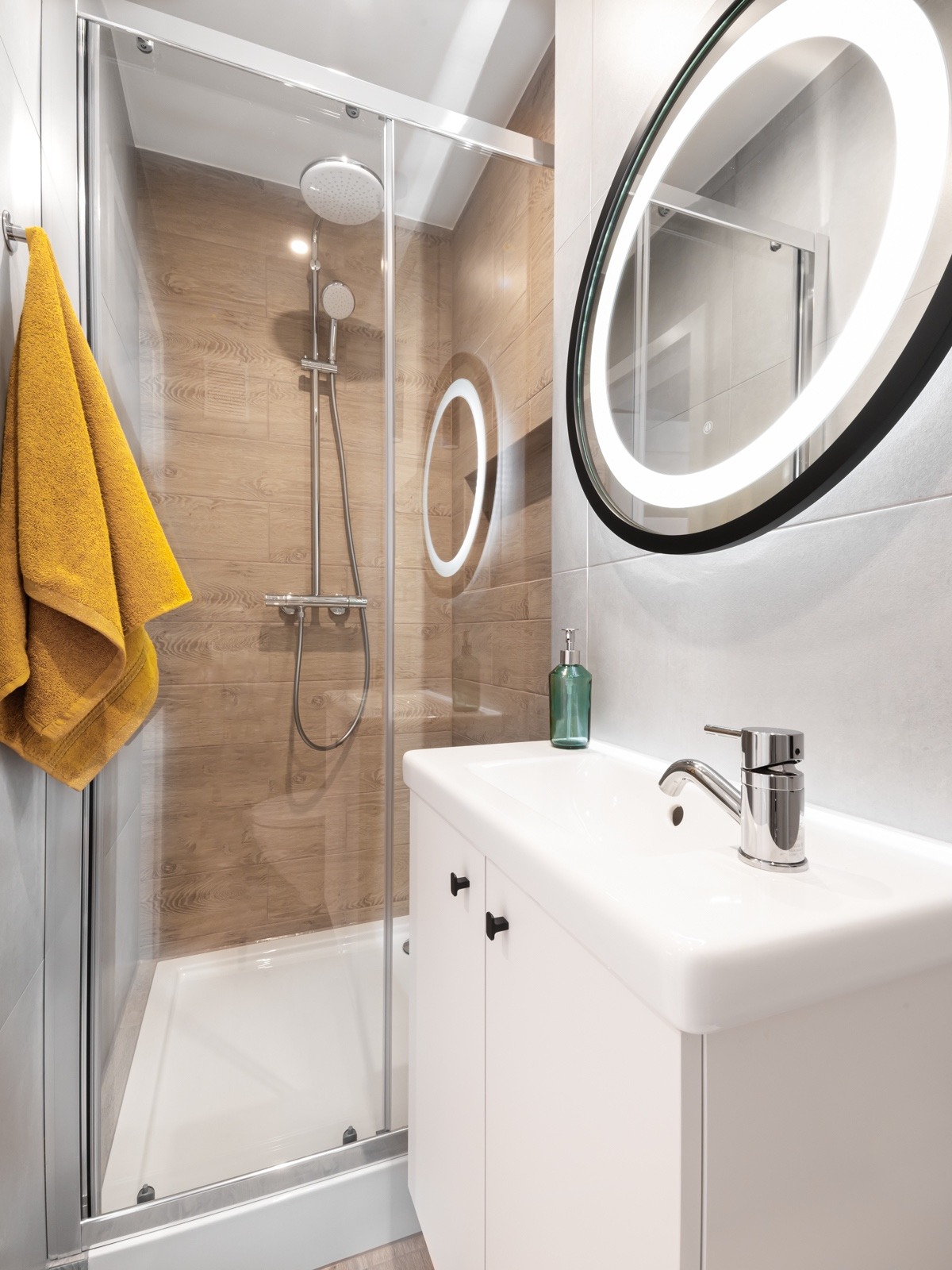 Nowoczesna łazienka z kabiną prysznicową, umywalką z szafką i okrągłym lustrem z podświetleniem LED. Na ścianach szare i drewnopodobne płytki. Na wieszaku żółty ręcznik.