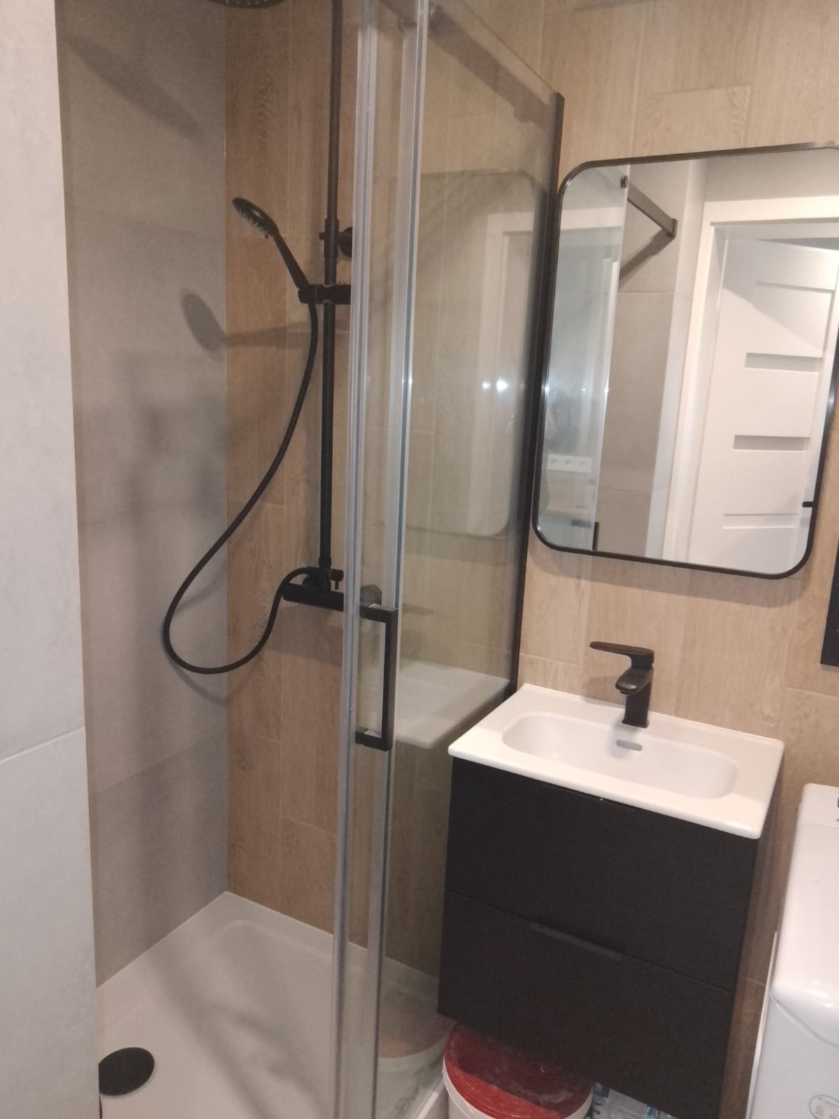 Nowoczesna łazienka z kabiną prysznicową typu walk-in, czarną armaturą i umywalką z szafką w kolorze ciemnego drewna, odbicie w lustrze ukazuje fragment białej ściany i drzwi.