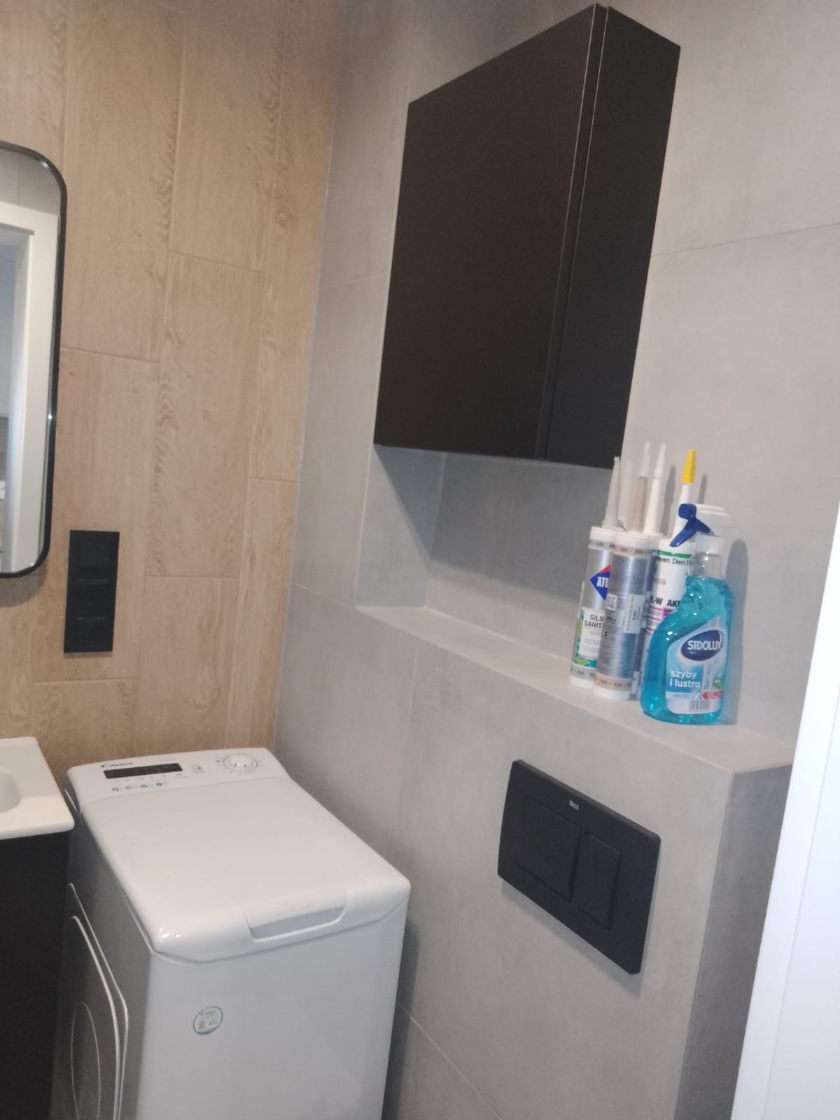 Nowoczesna łazienka z pralką Candy, wiszącą szafką w kolorze antracytu, płytkami imitującymi drewno i szare płytki, z widocznymi silikonami i płynem do szyb na półce.