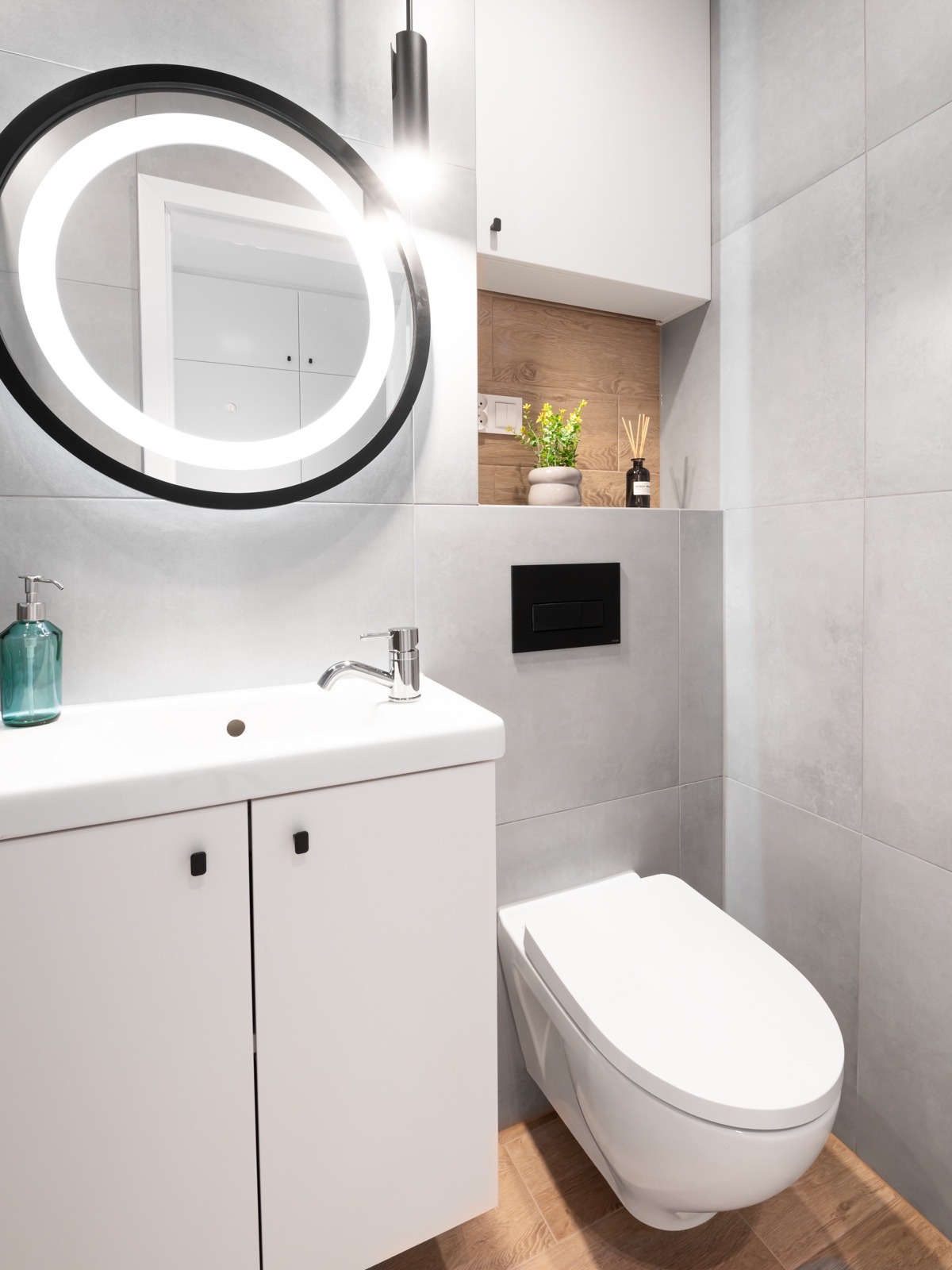 Nowoczesna łazienka z umywalką z szafką, podwieszaną toaletą i okrągłym lustrem z podświetleniem LED, wykończona szarymi płytkami ceramicznymi i drewnopodobną wnęką z rośliną i dyfuzorem zapachowym.
