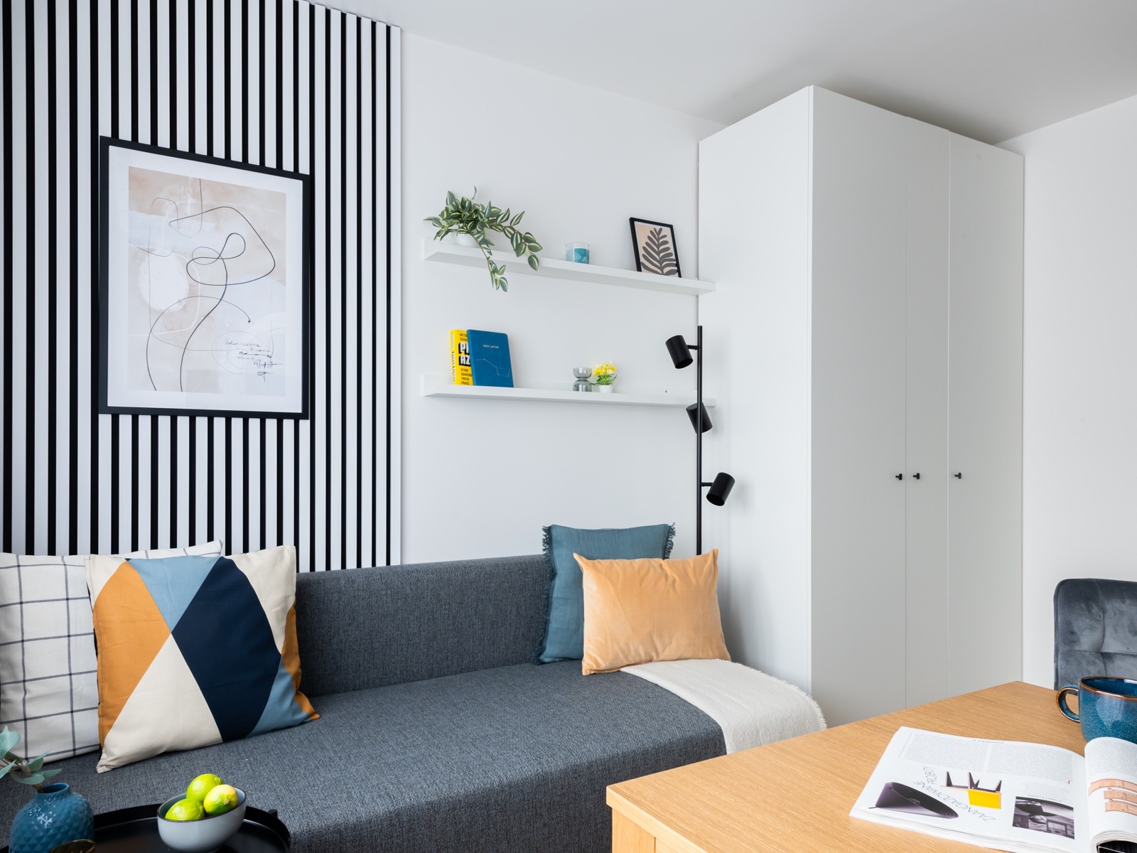 Jasne wnętrze pokoju dziennego z szarą sofą, kolorowymi poduszkami, półkami z dekoracjami, lampą podłogową i biurkiem z otwartą książką.