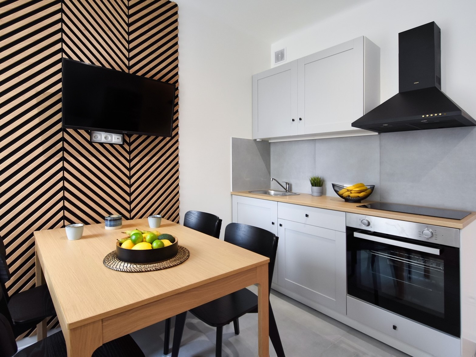 Nowoczesna kuchnia z jadalnią: drewniana ściana z telewizorem, jasne meble kuchenne, drewniany stół z owocami, czarne krzesła.