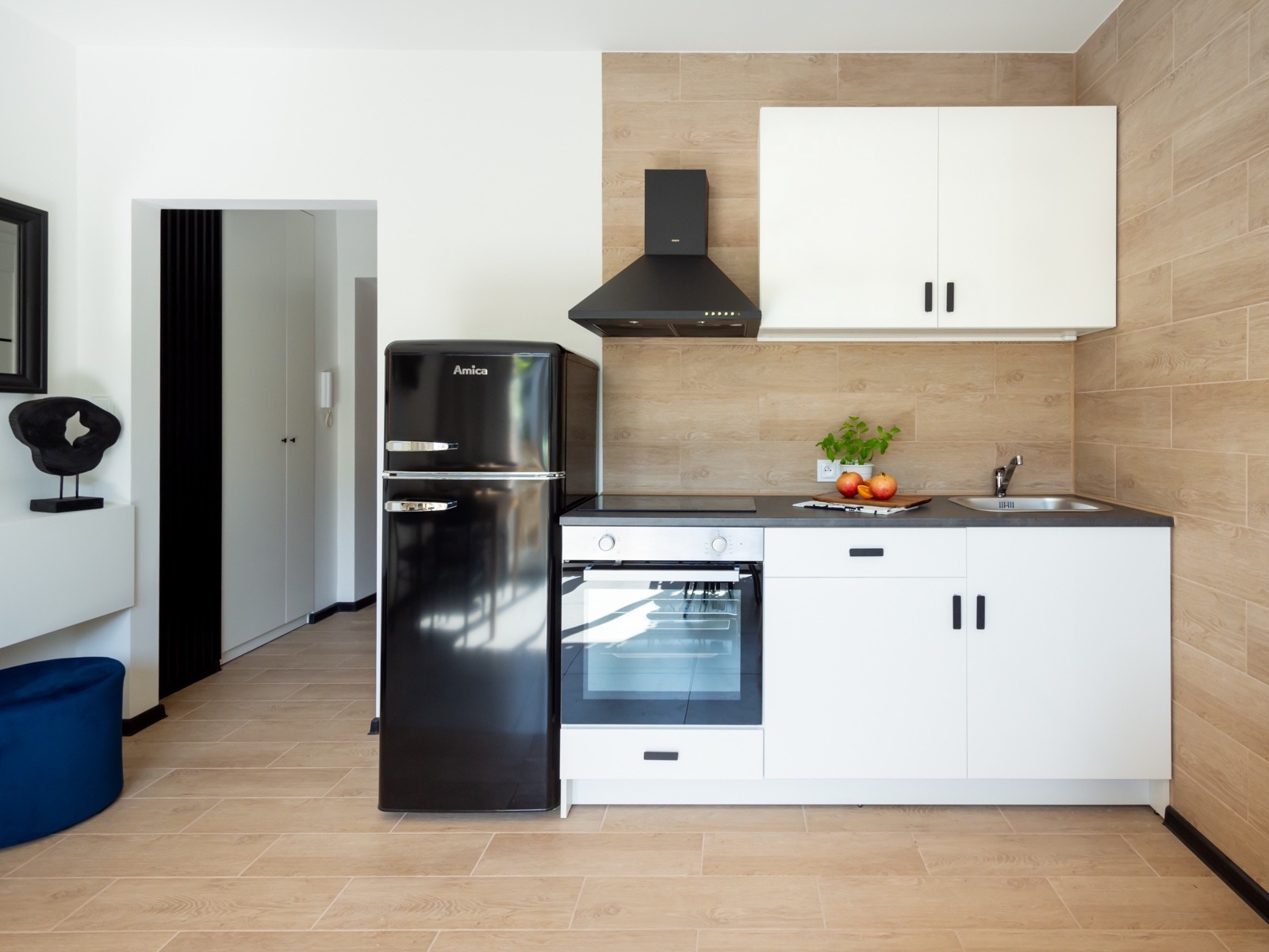 Nowoczesna kuchnia z czarną lodówką retro Amica, białymi szafkami i brązowymi płytkami imitującymi drewno, oświetlona naturalnym światłem w wykończonym mieszkaniu.