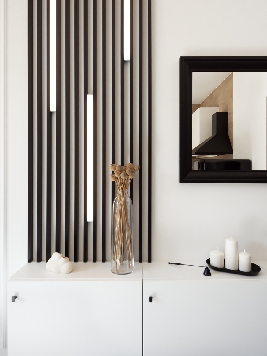 Nowoczesna aranżacja wnętrza: biała szafka z czarnymi uchwytami, dekoracyjna ścianka z pionowych listew z oświetleniem LED, lustro w czarnej ramie odbijające fragment kuchni z czarnym okapem, wazon...