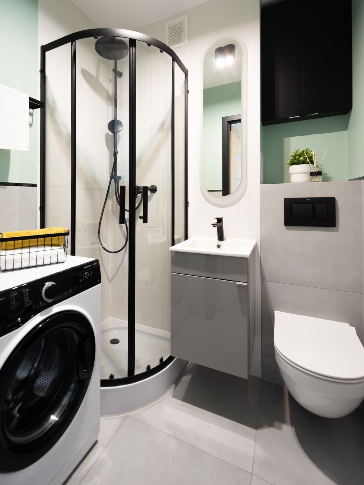 Nowoczesna łazienka w odcieniach szarości i bieli z akcentami czerni: kabina prysznicowa narożna z czarną ramą, pralka z czarnymi elementami, szara szafka pod umywalką, wisząca toaleta i podłużne...