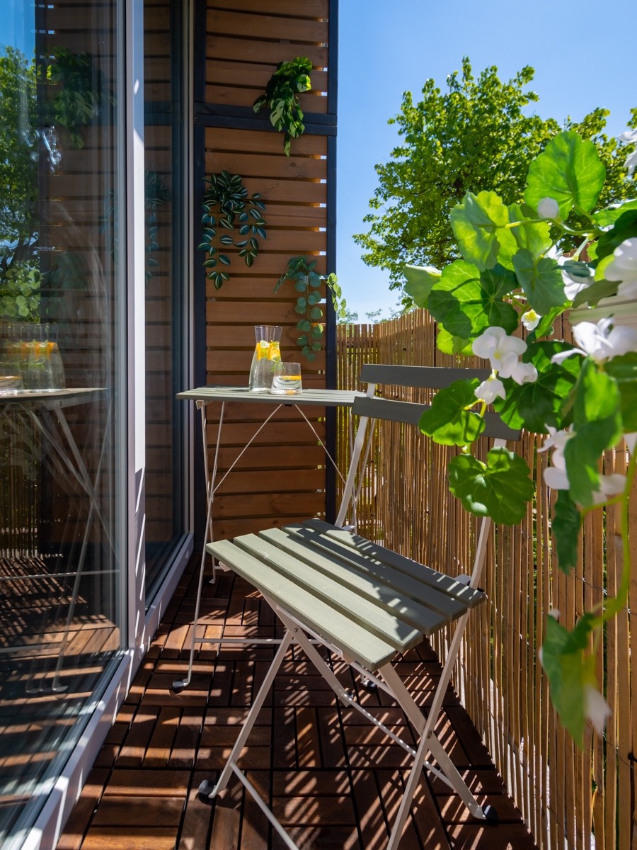 Zaciszny balkon z drewnianą podłogą, składanym stolikiem i krzesłem w odcieniach zieleni, dekorowany pnączami i kwiatami, z widokiem na zieleń drzew.