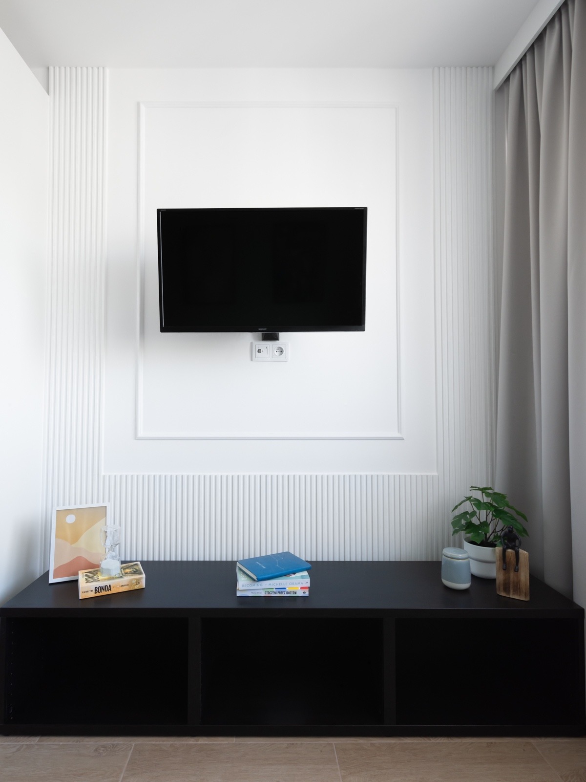 Nowoczesny salon z telewizorem na ścianie z dekoracyjnymi panelami, czarna szafka RTV z książkami i rośliną doniczkową, beżowe zasłony i podłoga z płytek.