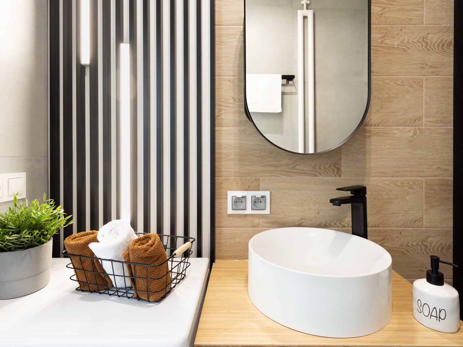 Nowoczesna łazienka z owalną umywalką nablatową, czarną armaturą i lustrem w czarnej ramie, akcentowana pionowymi panelami dekoracyjnymi w czarno-białe pasy oraz drewnopodobnymi płytkami.