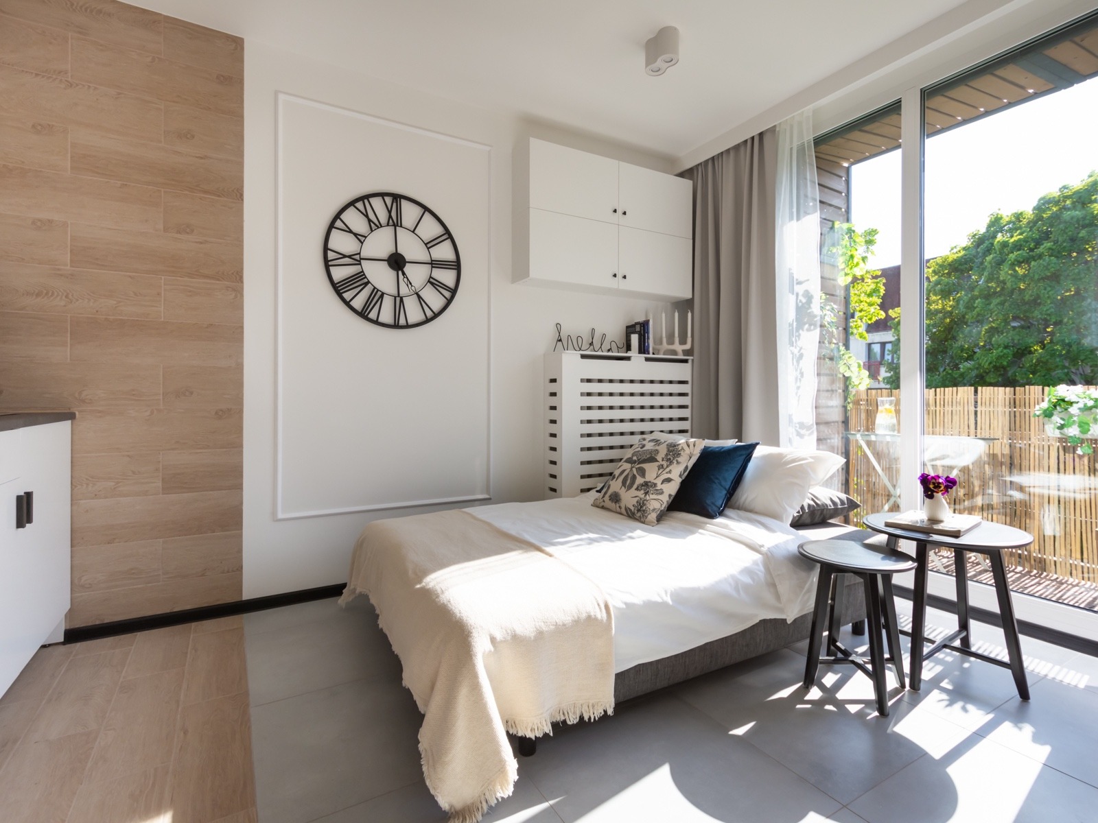Jasna sypialnia z wyjściem na balkon, drewnopodobna okładzina ściany, duży zegar na ścianie, szare panele podłogowe i minimalistyczne meble.