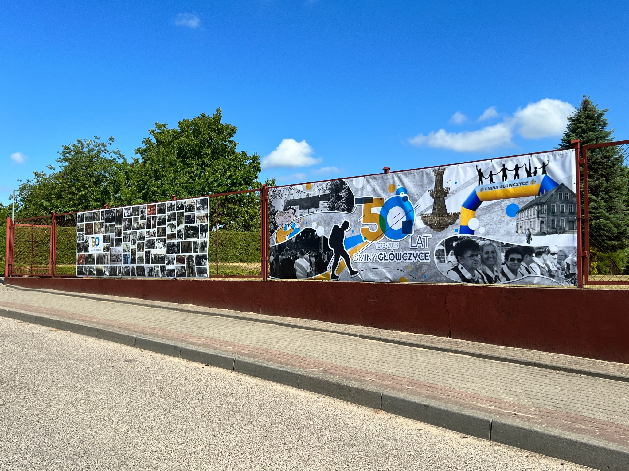 Dwa banery reklamowe na ogrodzeniu, upamiętniające 50-lecie Gminy Główczyce, z elementami graficznymi, zdjęciami i logotypami, widoczne z ulicy w słoneczny dzień.