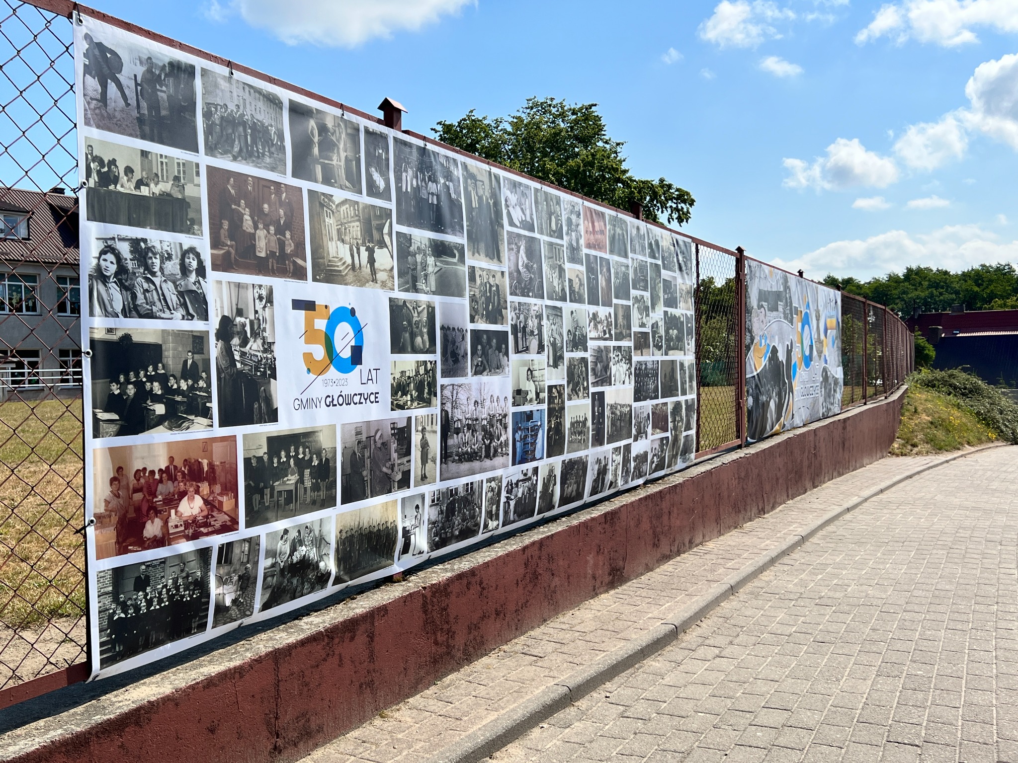 Baner z kolażem czarno-białych zdjęć, upamiętniający 50-lecie Gminy Główczyce, zawieszony na metalowym ogrodzeniu.
