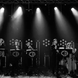 Czarno-białe zdjęcie zespołu muzycznego 'Commers Band' na scenie w trakcie występu, oświetlonego reflektorami punktowymi, z widocznymi instrumentami dętymi i wokalistami.