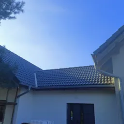 Nowy, ciemnoszary dach z blachodachówki modułowej, świeżo zamontowane rynny PCV, widok z dołu na tle błękitnego nieba, fragment drzewa iglastego w lewym górnym rogu.