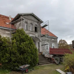 Remontowany budynek z rusztowaniem i stertą cegieł. Widoczny fragment dachu z czerwoną dach&oacute;wką i ciężar&oacute;wka. Prace dekarskie w toku, pochmurne niebo.