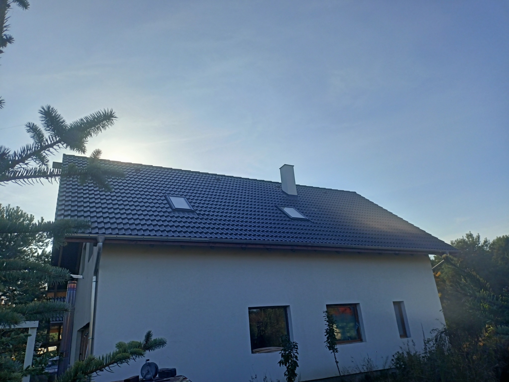 Dom jednorodzinny z ciemną dachówką, dwoma oknami dachowymi i białym kominem, widoczny z dołu pod kątem, z elementami zieleni na pierwszym planie i błękitnym niebem w tle.