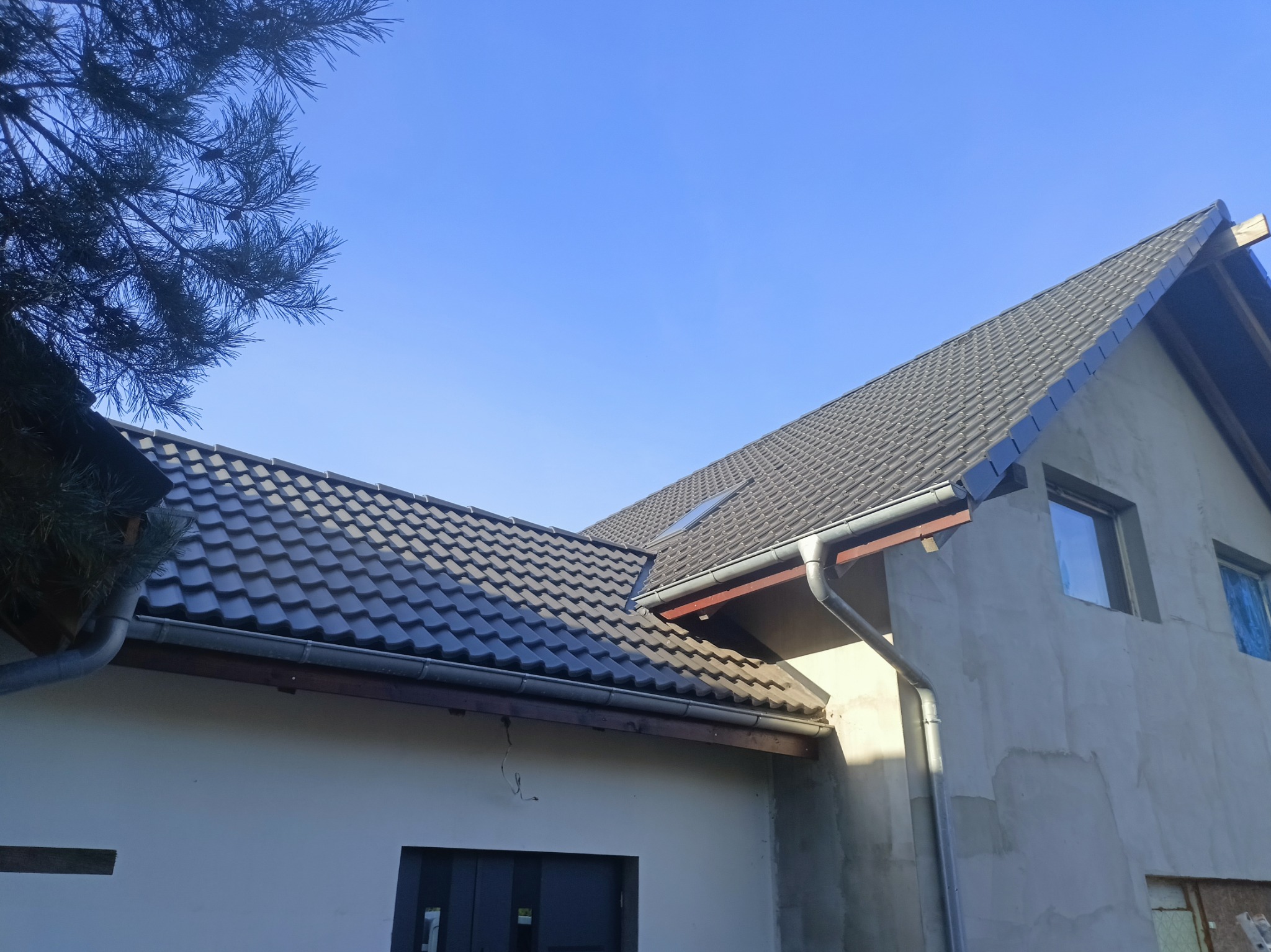 Dom w trakcie budowy z nowym, ciemnoszarym dachem z dachówki ceramicznej i zamontowanymi rynnami, widok z dołu, fragment drzewa iglastego w lewym górnym rogu.