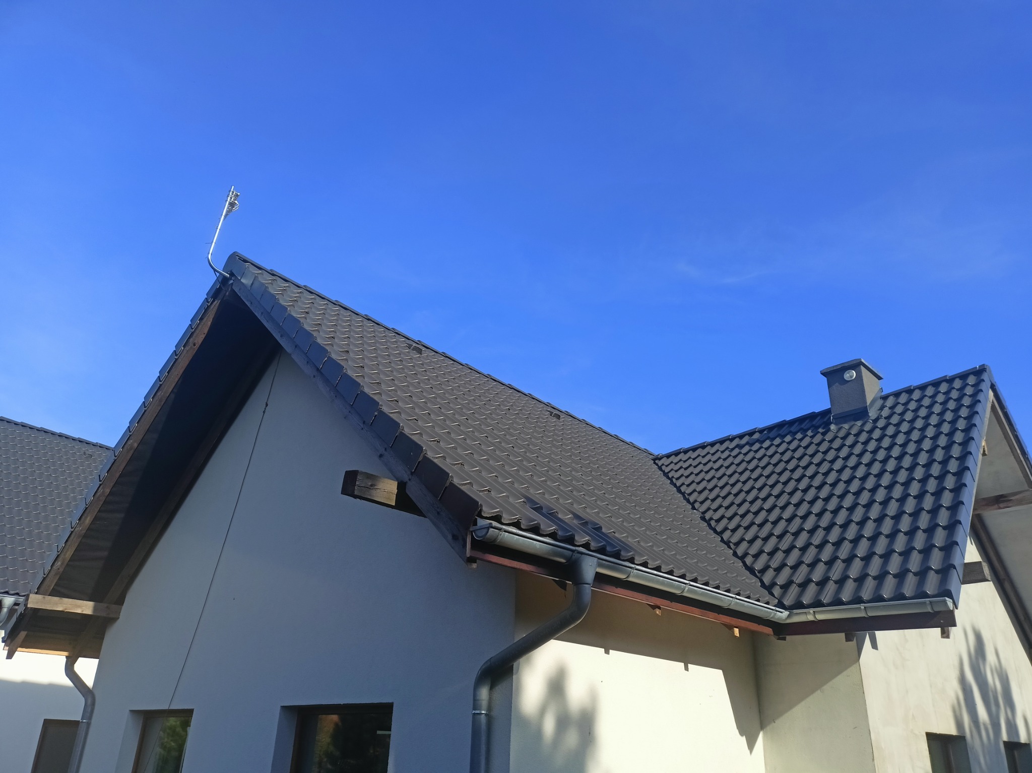 Nowy, ciemnoszary dach z blachodachówki na jasnym domu jednorodzinnym, widoczny komin i antena na dachu, błękitne niebo w tle.