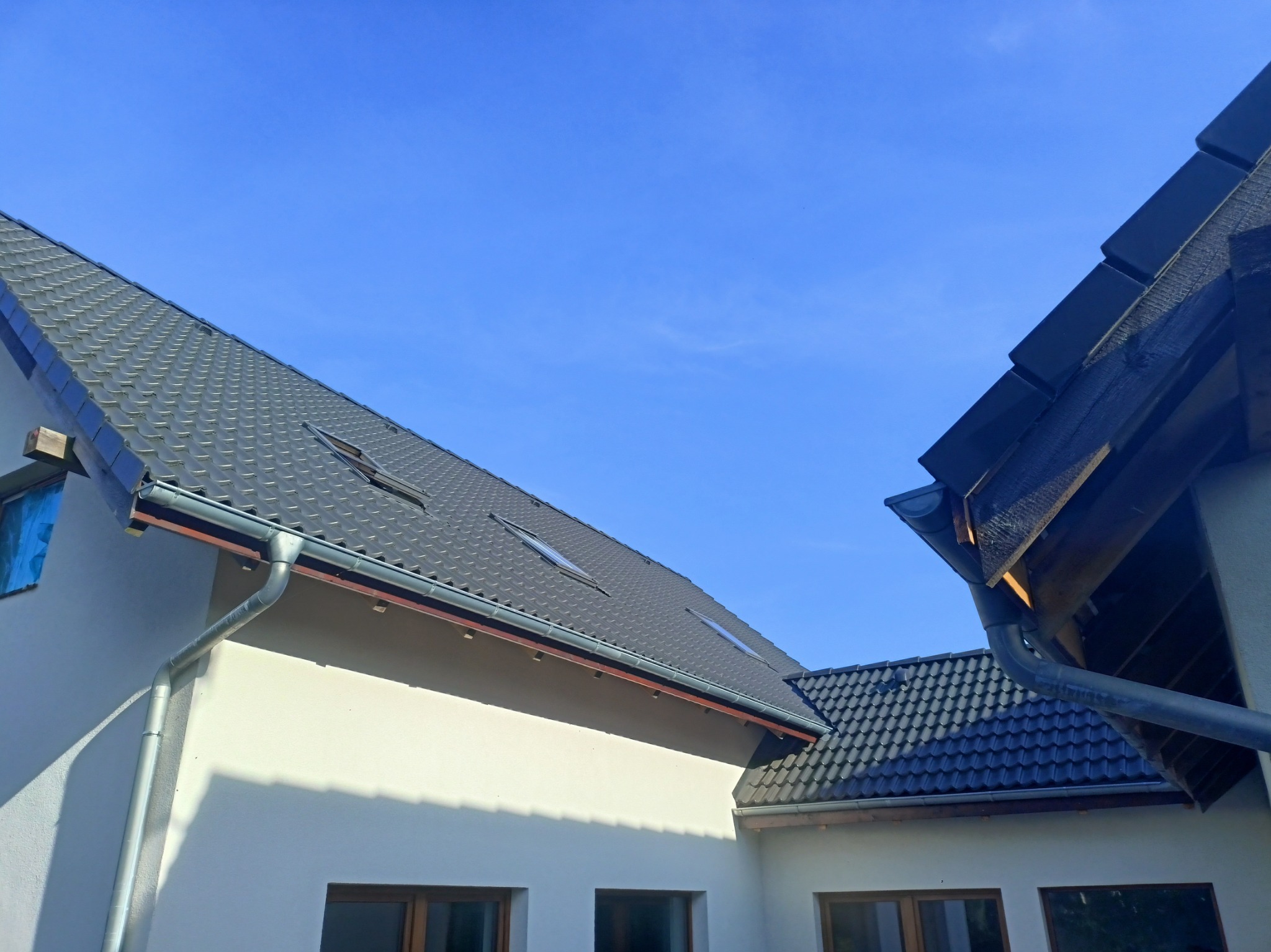 Nowoczesny dom z ciemnoszarą dachówką ceramiczną i zamontowanymi rynnami w kolorze grafitowym, widoczne okna dachowe i fragment elewacji w jasnym odcieniu.