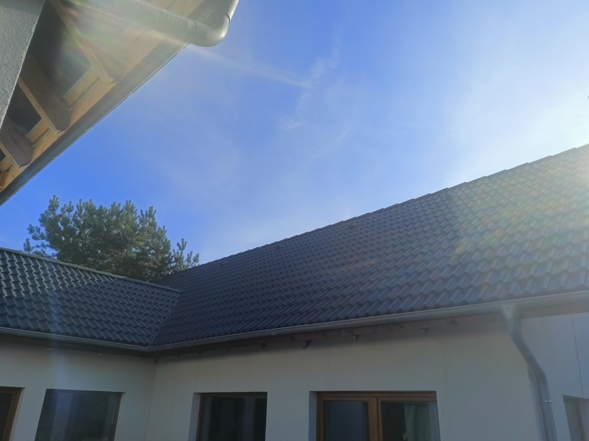 Ciemna dachówka ceramiczna na skośnym dachu domu jednorodzinnego, widok z dołu, rynna wzdłuż krawędzi dachu, fragment ściany budynku i drzewo iglaste w tle na tle błękitnego nieba.