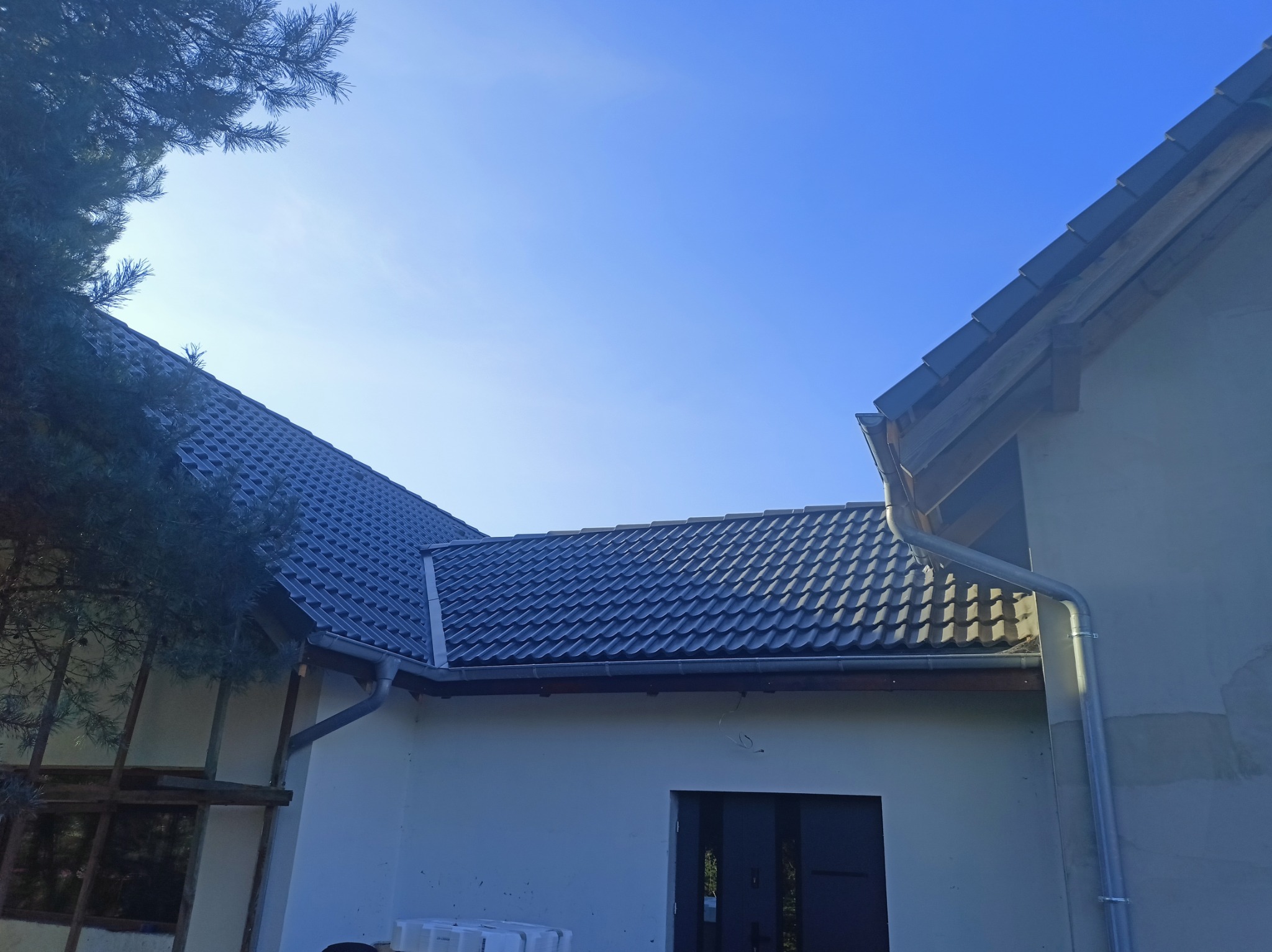 Nowy, ciemnoszary dach z blachodachówki modułowej, świeżo zamontowane rynny PCV, widok z dołu na tle błękitnego nieba, fragment drzewa iglastego w lewym górnym rogu.