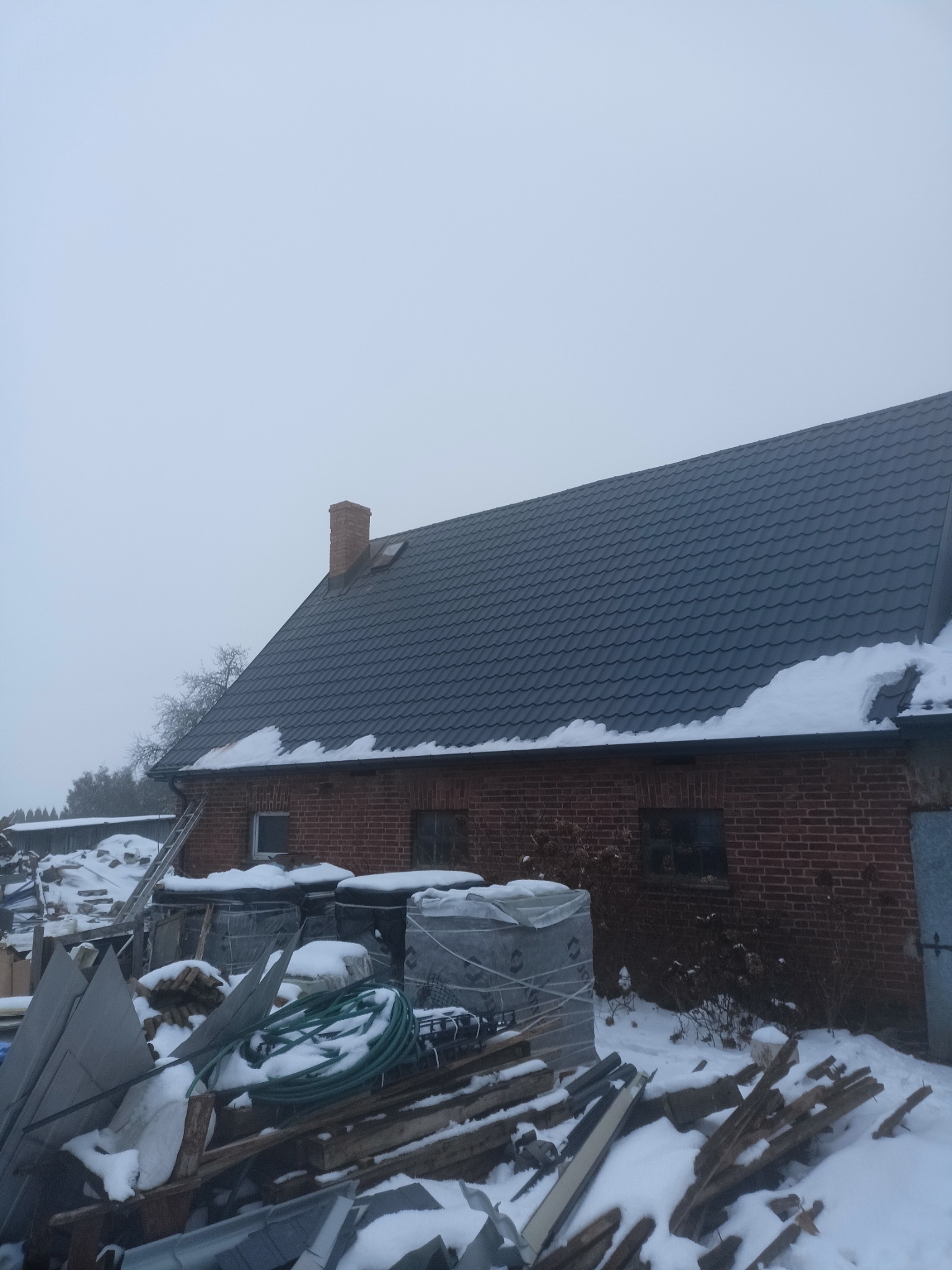 Dom z ciemnoszarą blachodachówką pokrytą śniegiem, widoczny komin i okno dachowe, na ziemi składowane materiały budowlane i narzędzia, pochmurna pogoda.