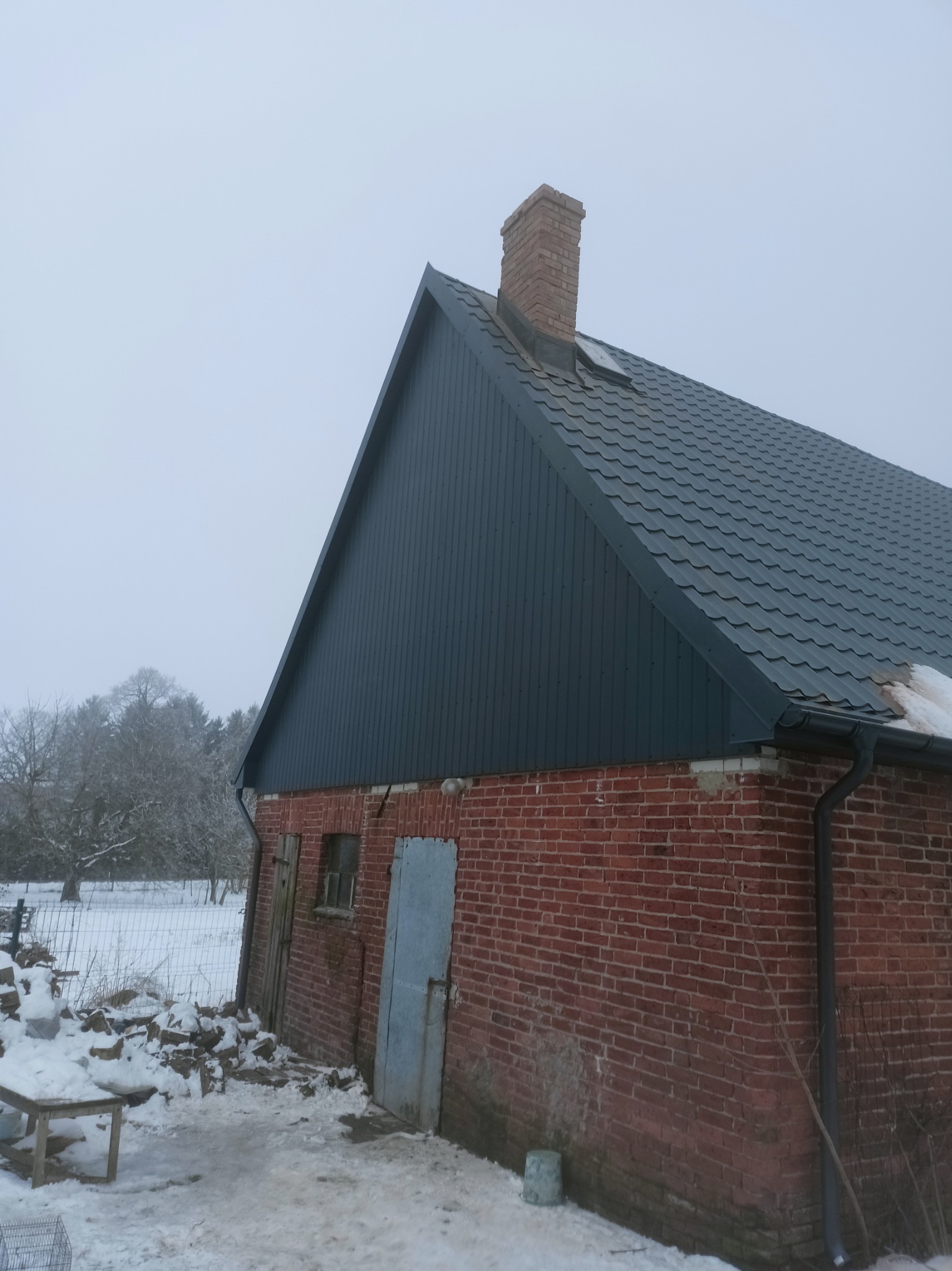 Ceglany budynek gospodarczy z nowym, grafitowym dachem z blachy, rynną i kominem w zimowej scenerii.