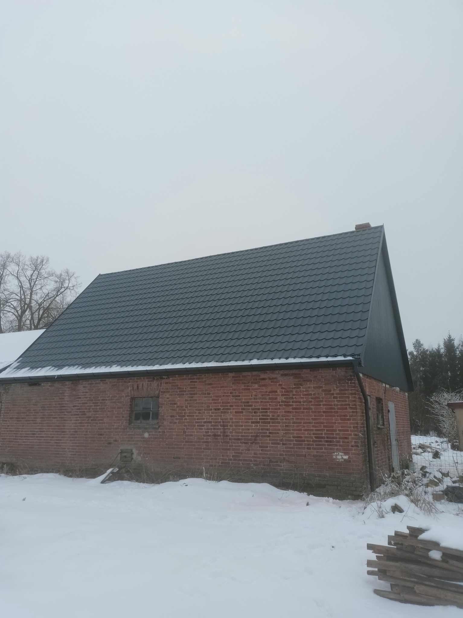 Ciemnoszary dach z blachy trapezowej na starym, ceglanym budynku gospodarczym w zimowej scenerii, śnieg leży na ziemi i częściowo na dachu.