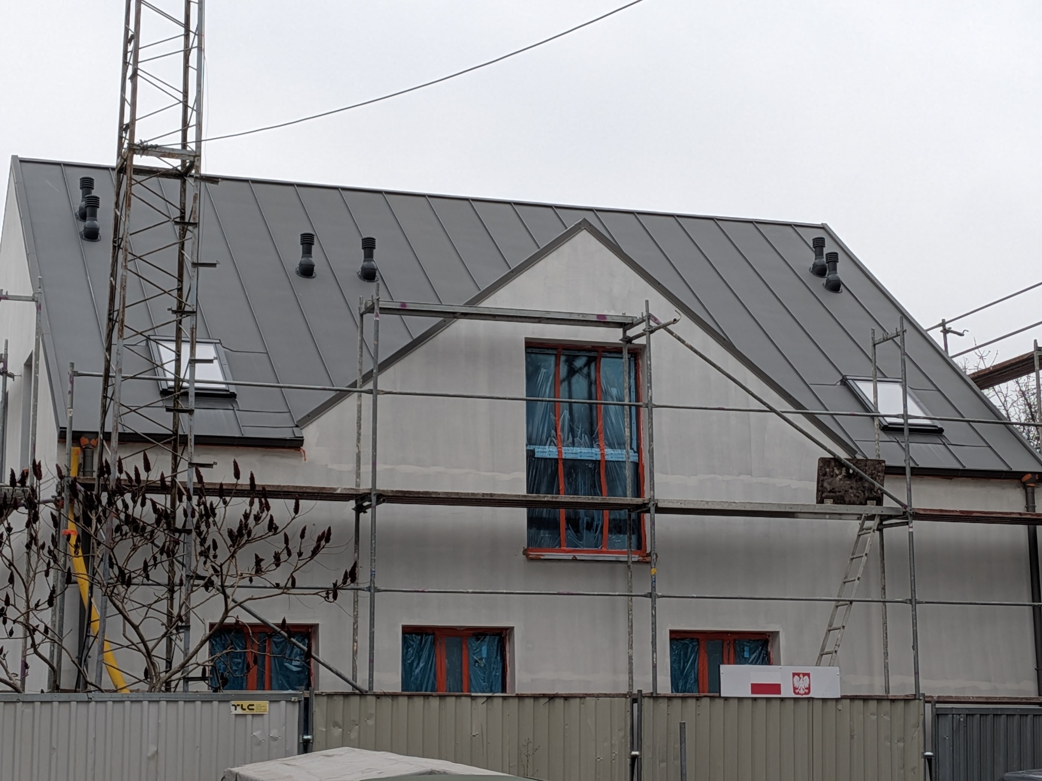 Nowoczesny dom w trakcie budowy z szarą blachą na dachu i rusztowaniami. Widoczne kominki wentylacyjne na dachu i okna zabezpieczone folią.