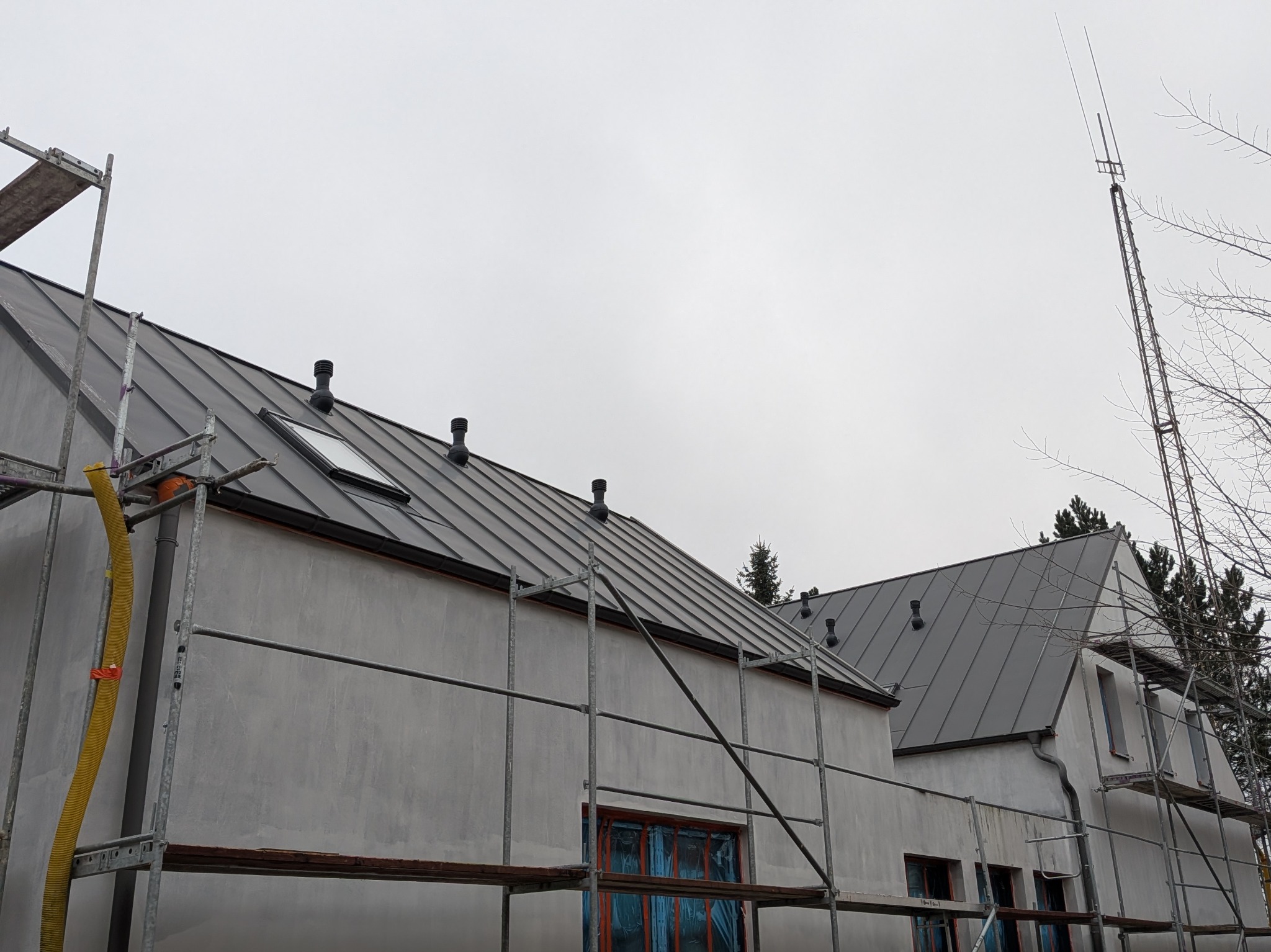 Szary dach z blachy na budynku w trakcie prac wykończeniowych, widoczne rusztowania i kominki wentylacyjne. Ujęcie z dołu, pochmurne niebo w tle.