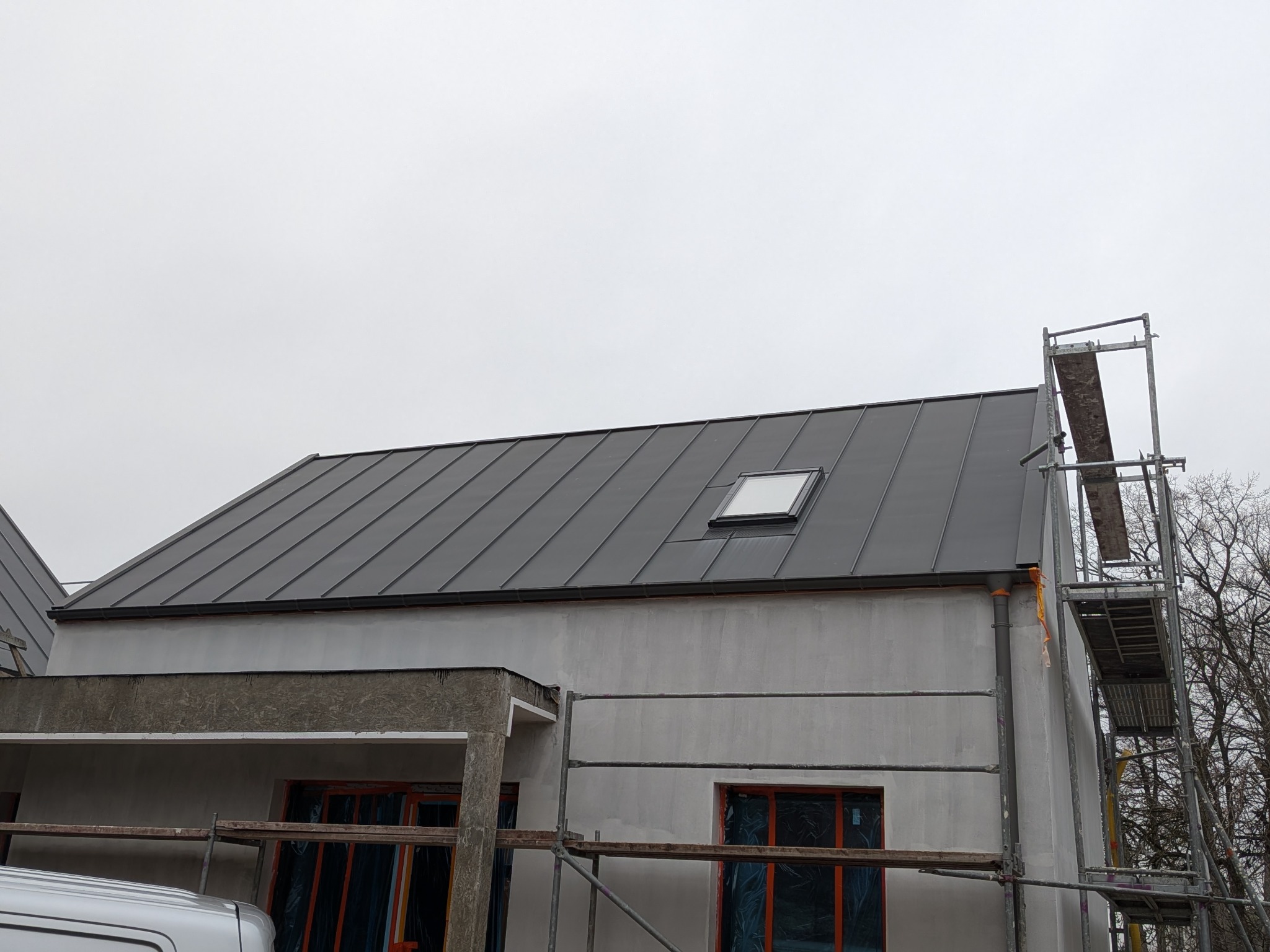 Nowoczesny dom w trakcie budowy z szarym dachem z blachy na rąbek stojący i oknem dachowym. Widoczne rusztowania i fragment samochodu dostawczego.