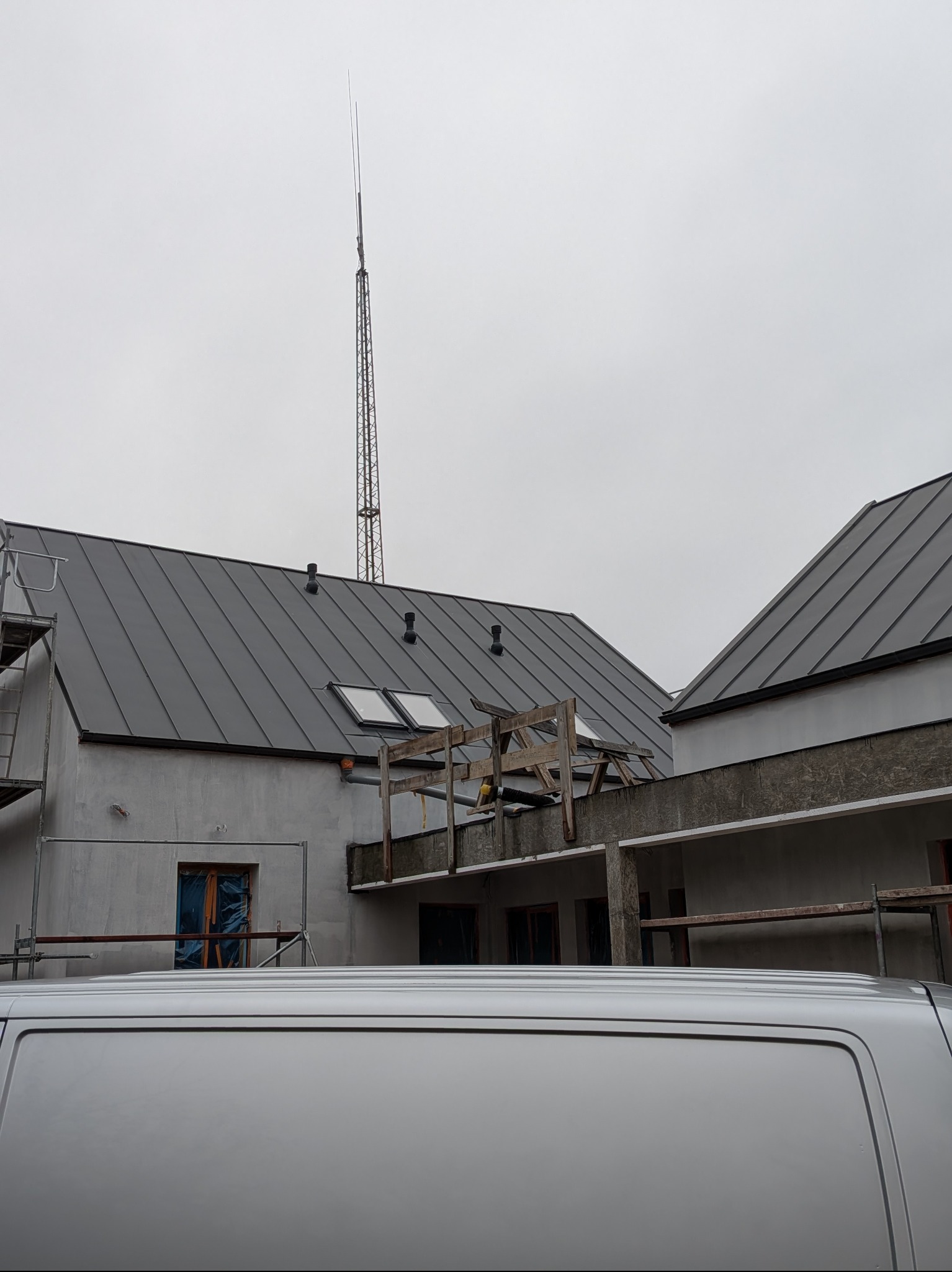 Szare dachy budynków w trakcie budowy, widoczne okna dachowe i rusztowania. Na pierwszym planie fragment białego samochodu dostawczego. W tle wysoka wieża telekomunikacyjna.