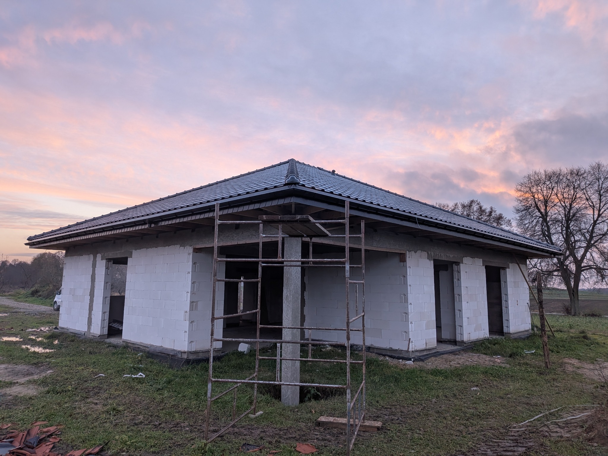 Niedokończony dom z białych bloczków i ciemnym dachem na tle pastelowego nieba. Widoczne rusztowanie i fragmenty dachówek. Ujęcie z poziomu gruntu.