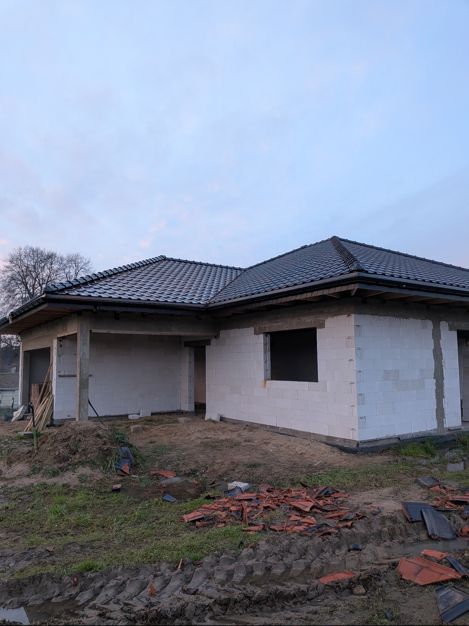Dom w trakcie budowy z częściowo ukończonym dachem z ciemnej dachówki. Widoczne ściany z betonu komórkowego i fragmenty porozrzucanej dachówki na ziemi.