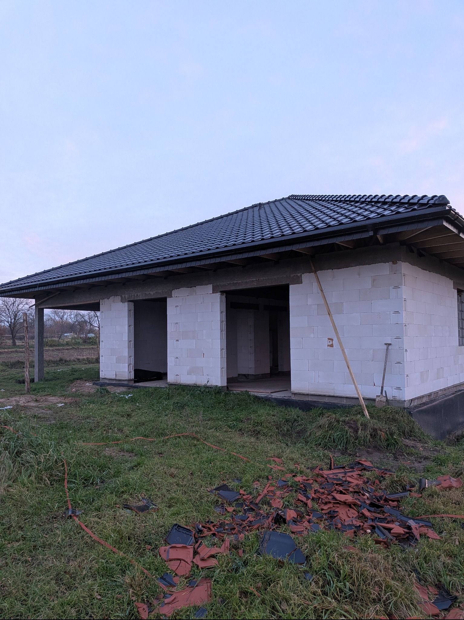 Budynek w trakcie budowy z ciemną dachówką, widoczne białe bloczki konstrukcyjne i resztki starej dachówki na trawie. Szpadel oparty o ścianę.