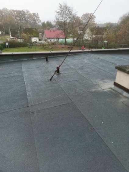 Czarny, płaski dach pokryty papą z widocznym odgromem i elementami wentylacji na tle zabudowań mieszkalnych w pochmurny dzień. Widoczne łączenia papy.