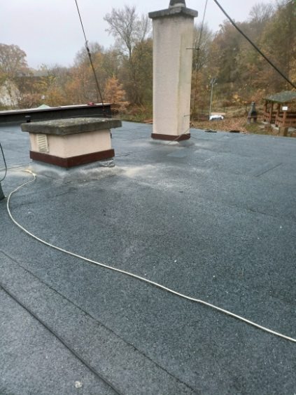 Pokrycie dachu papą termozgrzewalną z kominem i wentylacją. Widoczne ślady posypki mineralnej. Tło stanowią drzewa i altana.