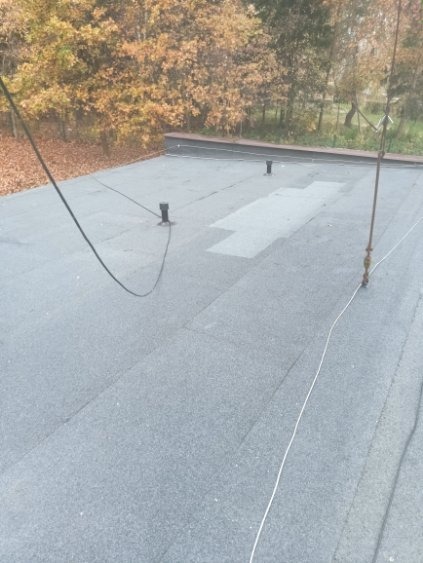 Płaski dach pokryty papą z widocznymi kominkami wentylacyjnymi i łatą. W tle drzewa z jesiennymi liśćmi. Widok z góry.