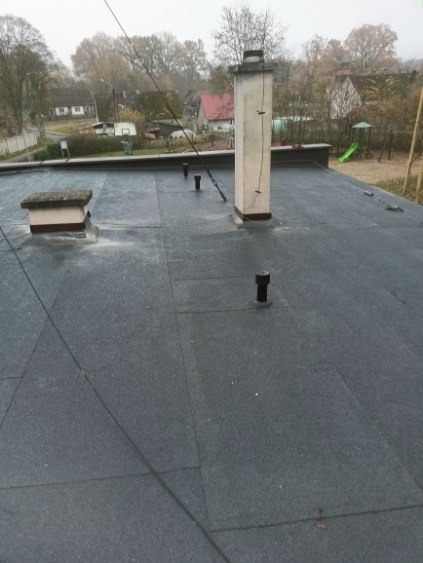 Czarny, płaski dach pokryty papą z kominem i odpowietrznikami na tle zabudowy jednorodzinnej i drzew w pochmurny dzień. Widoczne łączenia papy.