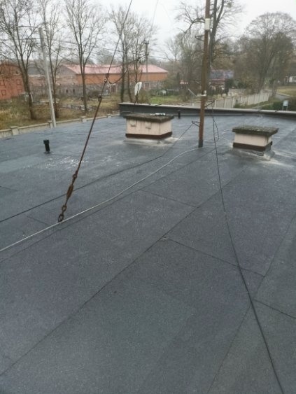 Czarny, płaski dach pokryty papą termozgrzewalną z dwoma kominami wentylacyjnymi. W tle budynki i drzewa. Widoczne elementy instalacji odgromowej.