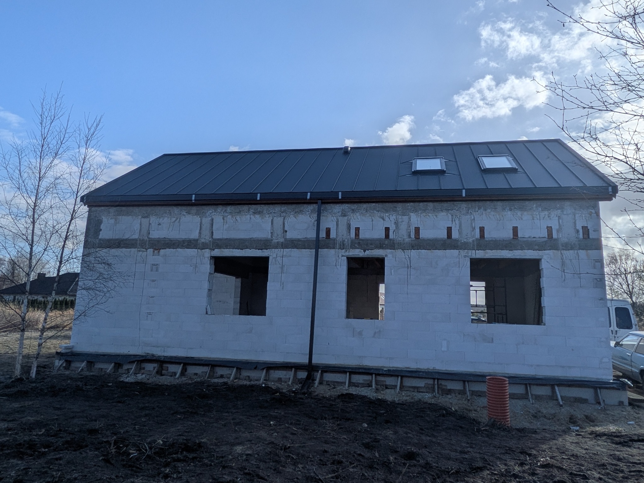 Dom w trakcie budowy z ciemnym dachem i oknami dachowymi. Widoczne bloczki konstrukcyjne i przygotowane otwory okienne. Niebo z chmurami w tle.