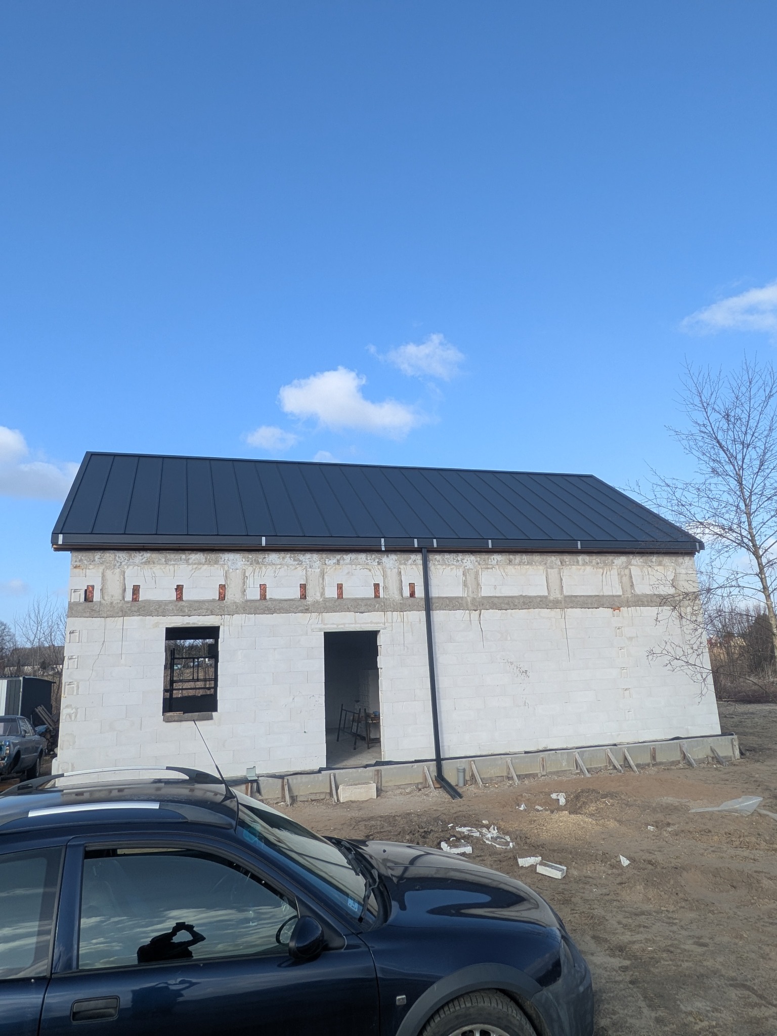 Nowy, grafitowy dach z blachy na surowym budynku z betonu komórkowego w słoneczny dzień. Widoczne rynny i obróbki blacharskie. W tle błękitne niebo.