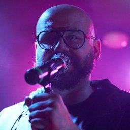 Szymon Skibiński - Łysy mężczyzna w okularach śpiewa do mikrofonu na scenie oświetlonej fioletowym i niebieskim światłem.