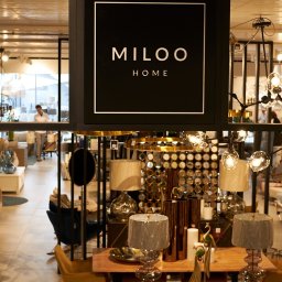 Szymon Skibiński - Wnętrze sklepu z wyposażeniem wnętrz, widoczne lampy, krzesła, dekoracje i logo 'MILOO HOME' na czarnym szyldzie.