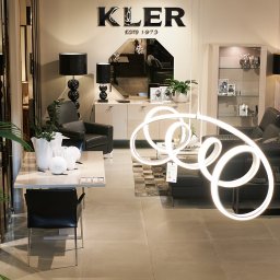 Szymon Skibiński - Salon meblowy Kler: czarny skórzany narożnik, stół z krzesłami, dekoracyjne lampy kuliste i modernistyczna lampa wisząca w kształcie spirali, lustro nad komodą, rośliny doniczkowe.