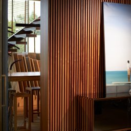 Szymon Skibiński - Wnętrze z drewnianą ścianą z listew, widoczne schody i obraz z motywem morskim, oświetlenie górne.