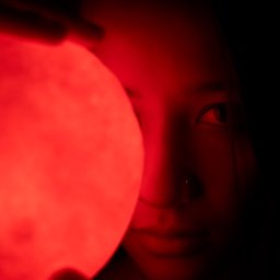 Szymon Skibiński - Portret kobiety oświetlonej czerwonym światłem z okrągłej lampy, z widocznym kolczykiem w nosie, na ciemnym tle.