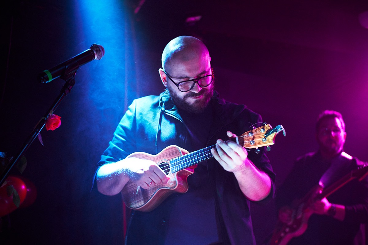 Muzyk z brodą i okularach gra na ukulele podczas występu na scenie w Krakowie, oświetlonej niebieskim i różowym światłem, z mikrofonem i gitarzystą w tle.
